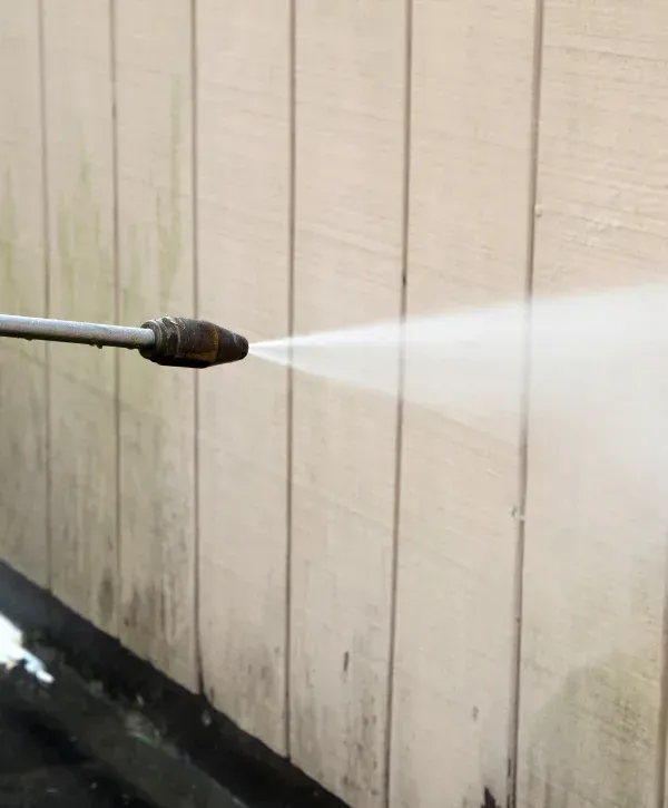 A close-up of a pressure washer nozzle spraying water onto dirty white house siding, cleaning the grime.
