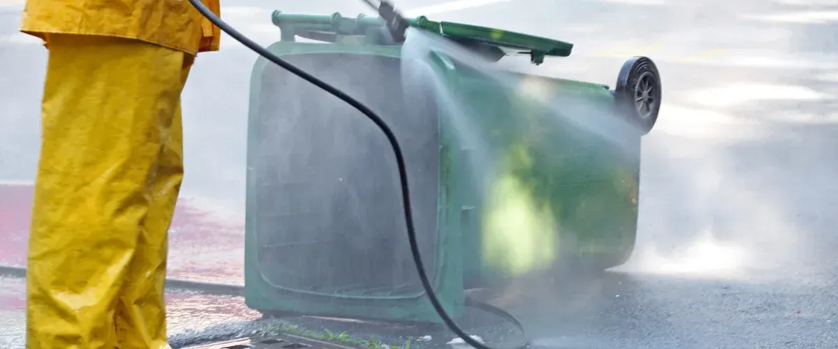 A person in yellow rain gear power washing a green garbage bin lying on its side on asphalt.