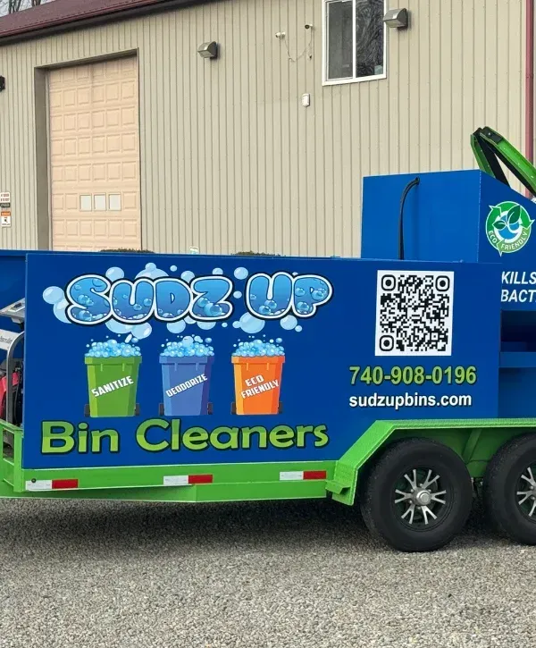 A full view of the Suds Up Bin Cleaners trailer, bright blue and green, attached to a white pickup truck on a gravel lot under a cloudy sky.