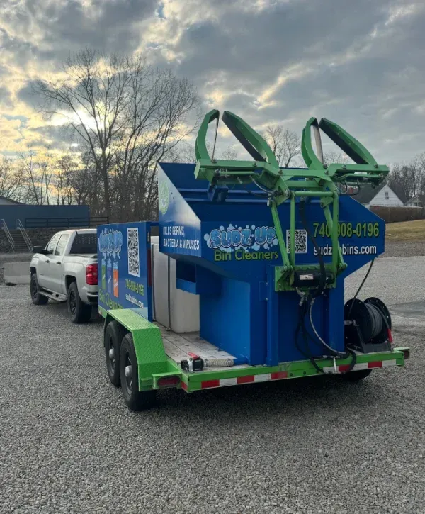 Side view of a blue and green 'Sudz Up Bin Cleaners' trailer with logo, QR code, phone number, and website.