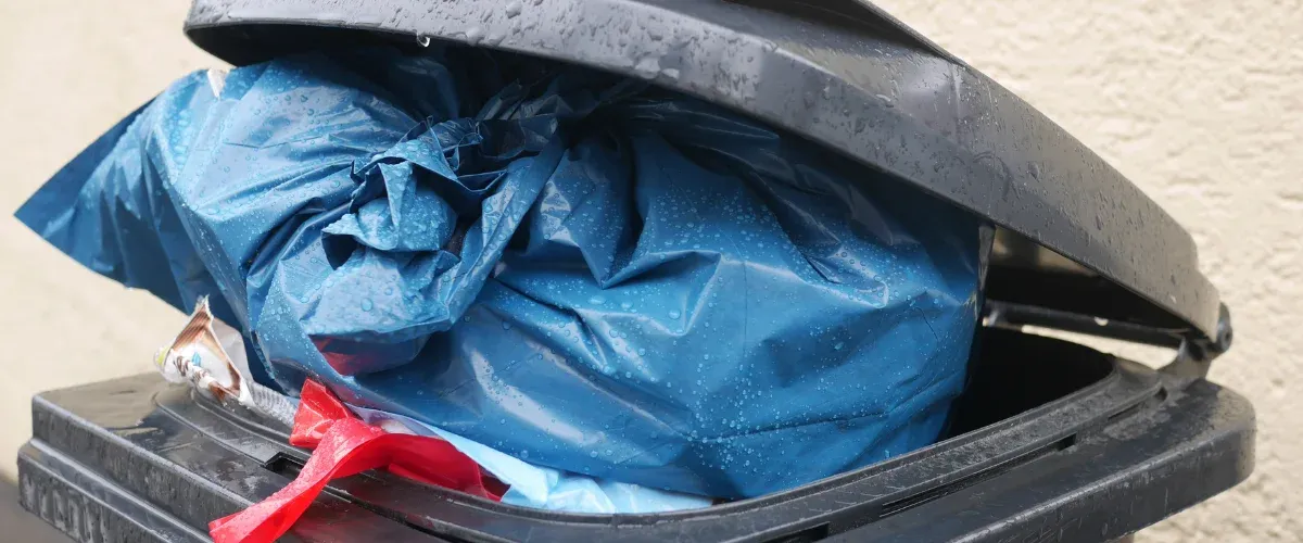 A close-up of a dark gray trash bin with an overflowing, rain-soaked blue garbage bag and a slightly ajar lid.