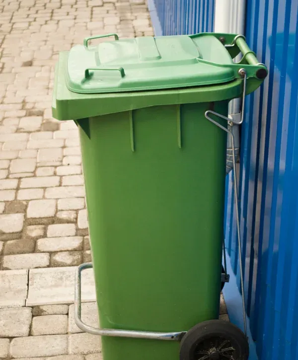 A bright green wheeled trash bin with a partially open lid stands on cobblestones next to a blue corrugated wall.