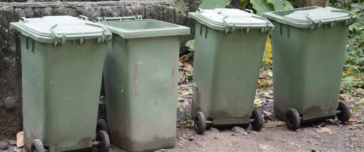 Four dirty green plastic waste bins, one with an open lid, lined up outdoors with foliage in the background.