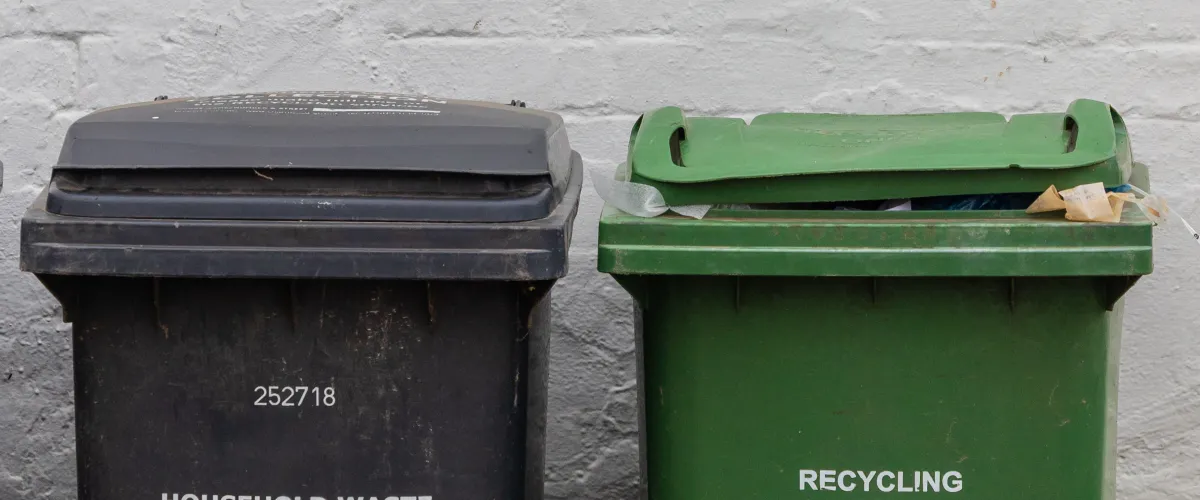 A black household waste bin next to a green recycling bin with a partially open lid against a white wall.