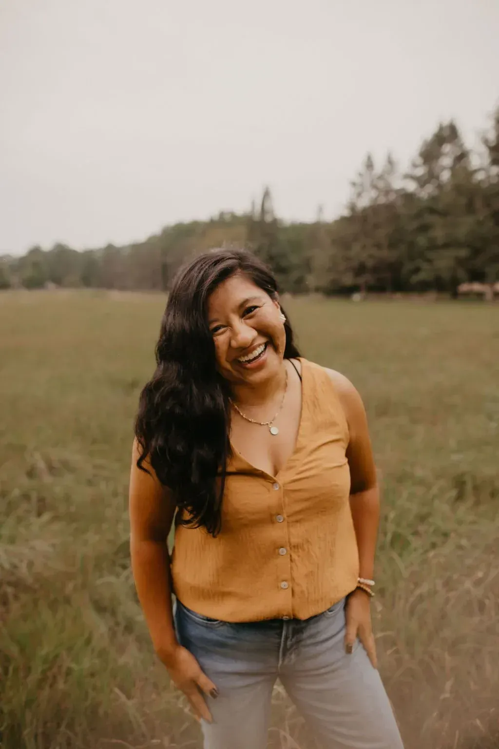 A smiling woman in her late 30s with long dark hair, wearing a flowing white dress, standing barefoot in a sun-dappled garden. She holds a single white feather, radiating warmth and wisdom. The background is lush and green, evoking peace and authenticity.