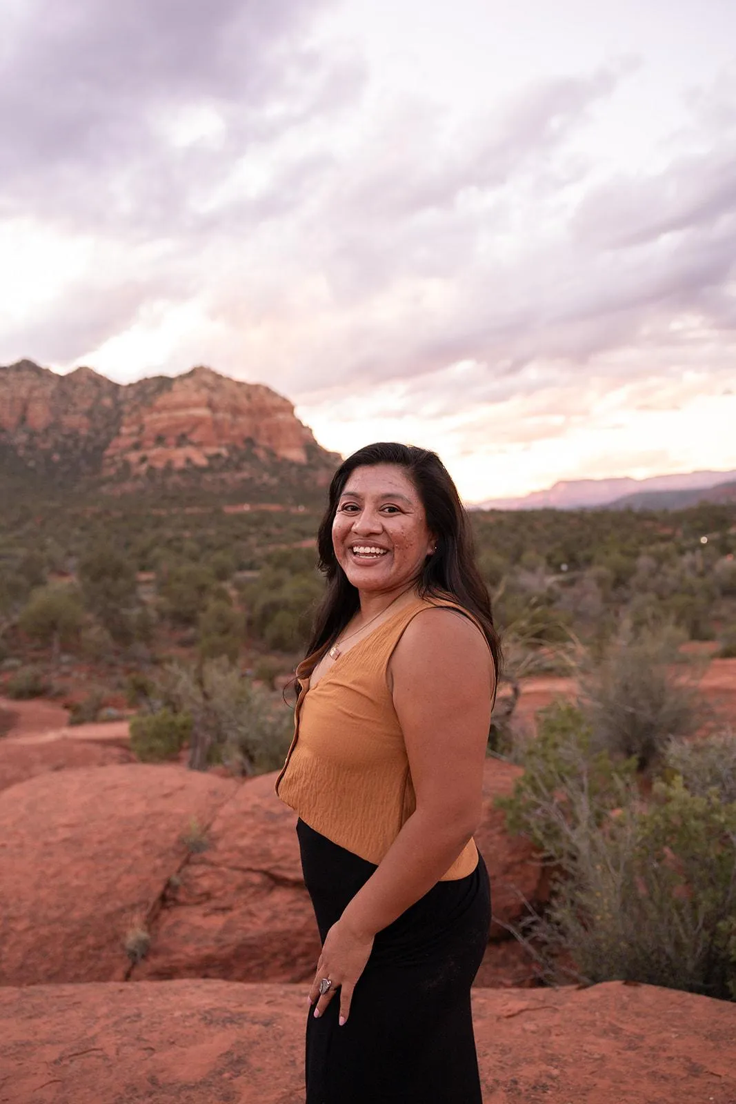 A smiling woman in her late 30s with long dark hair, wearing a flowing white dress, standing barefoot in a sun-dappled garden. She holds a single white feather, radiating warmth and wisdom. The background is lush and green, evoking peace and authenticity.