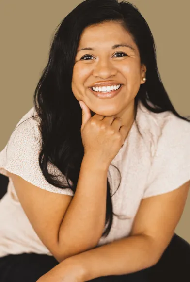 A smiling woman in her late 30s with long dark hair, wearing a flowing white dress, standing barefoot in a sun-dappled garden. She holds a single white feather, radiating warmth and wisdom. The background is lush and green, evoking peace and authenticity.