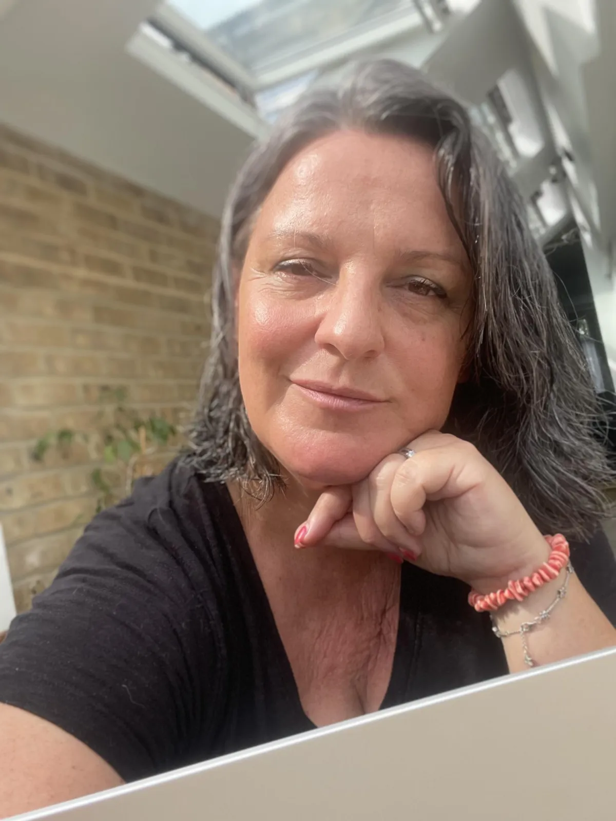 Atty in a sunny kitchen, smilig over a laptop, pral bracelet and nails, brick wll behind. A relaxed, friendly and uplifting vibe.