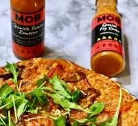 Two bottles of MOB Sauce next to a pizza with arugula.