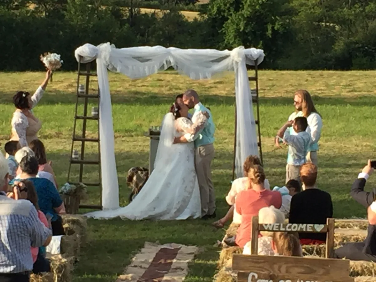 A bride and groom underneath an arch wrapped in tulle, kissing and surrounded by wedding guests.