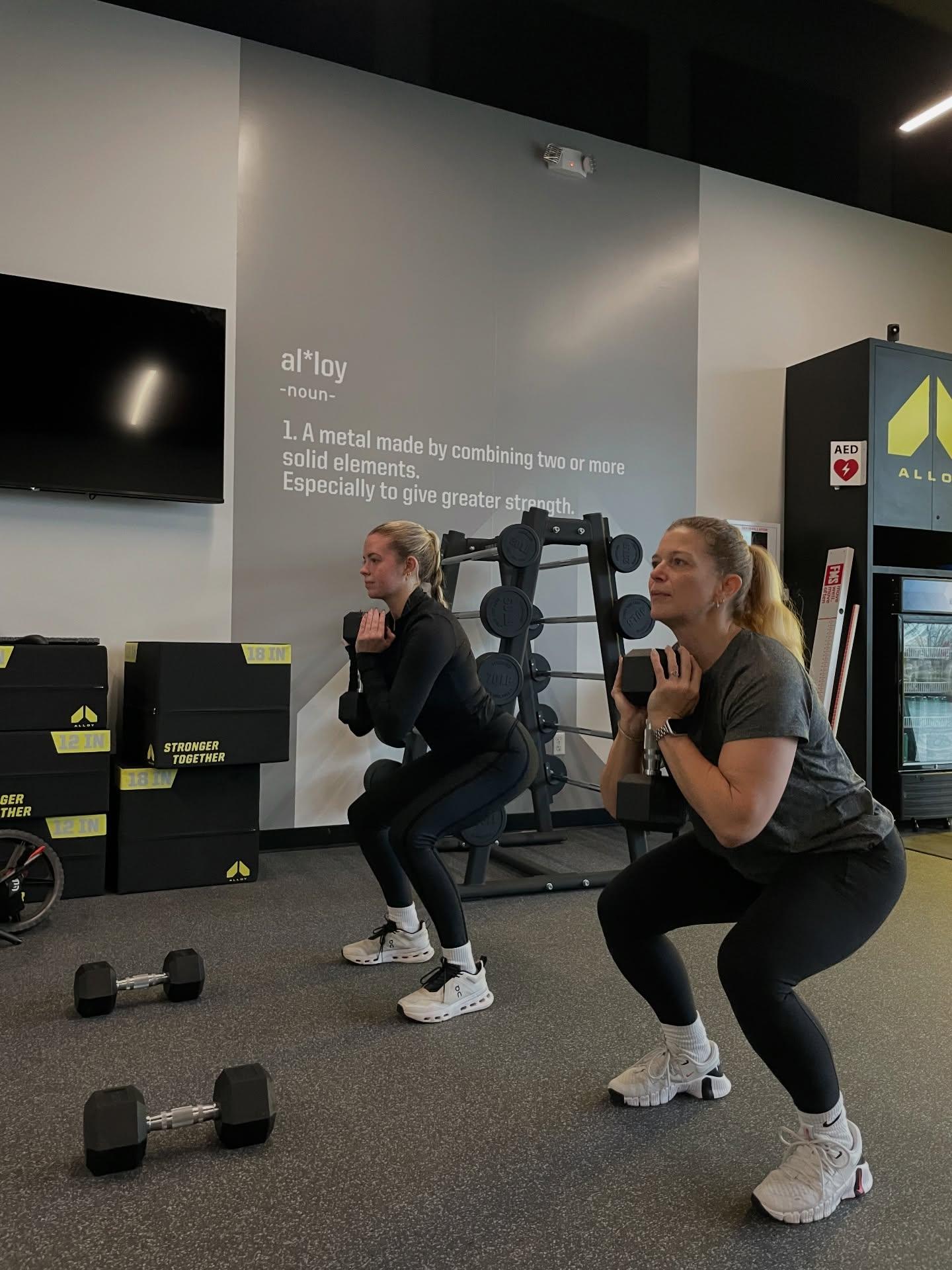 Two women performing kettlebell and dumbbell overhead carries during small group functional strength training at Alloy Personal Training Cedar Hills Crossing.