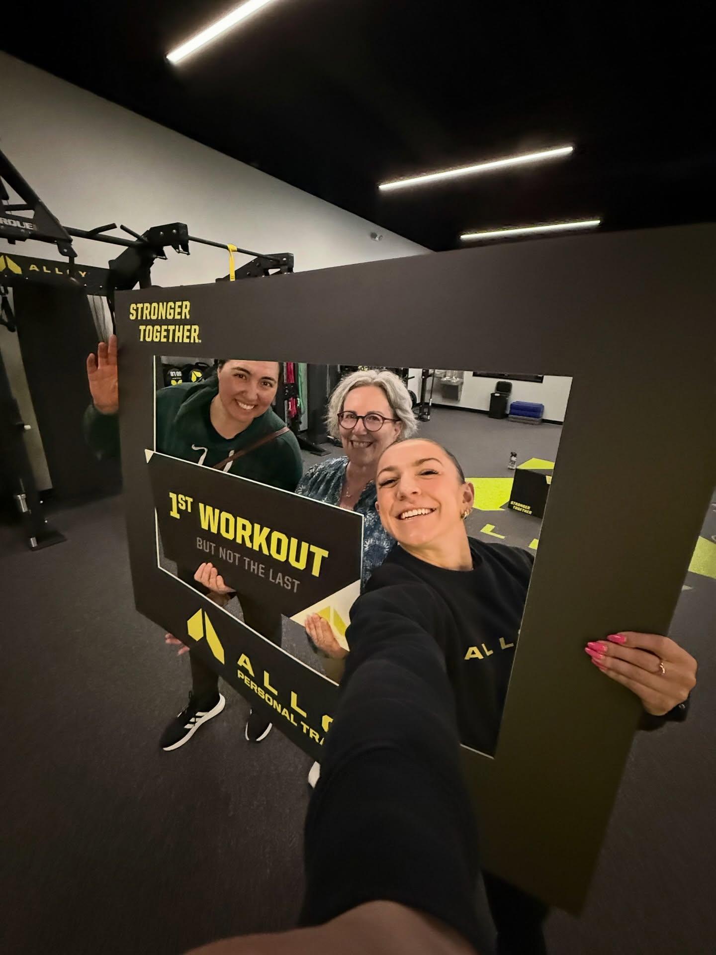 Alloy Personal Training Cedar Hills Crossing members holding 25 and 150 workout milestone signs in a small group personal training studio designed for adults over 40.