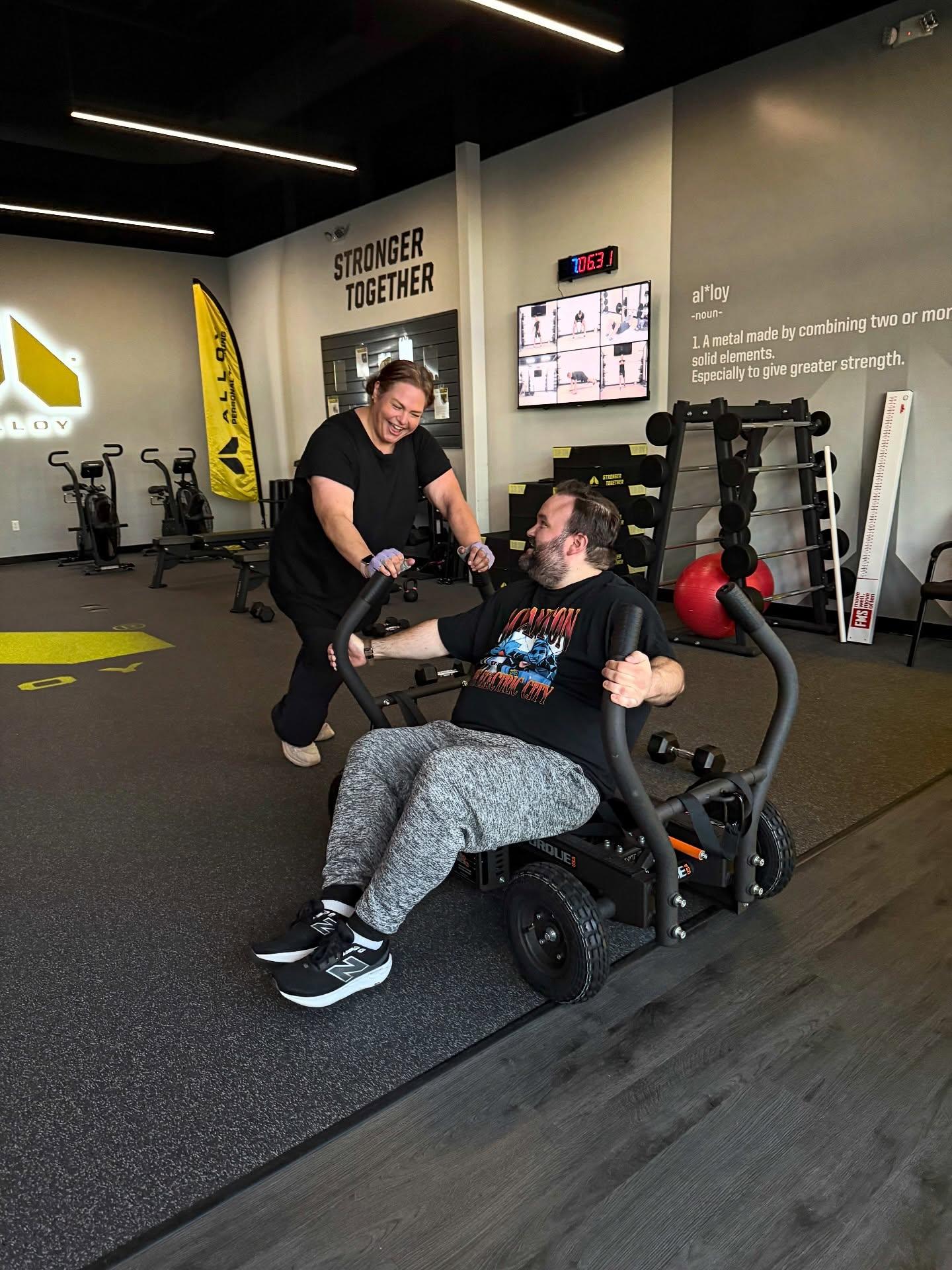 A personal trainer at Alloy Personal Training Cedar Hills Crossing assists a client with a sled push, part of a functional strength training session, in a well-equipped training space. This exercise improves power and mobility while engaging full-body muscles.