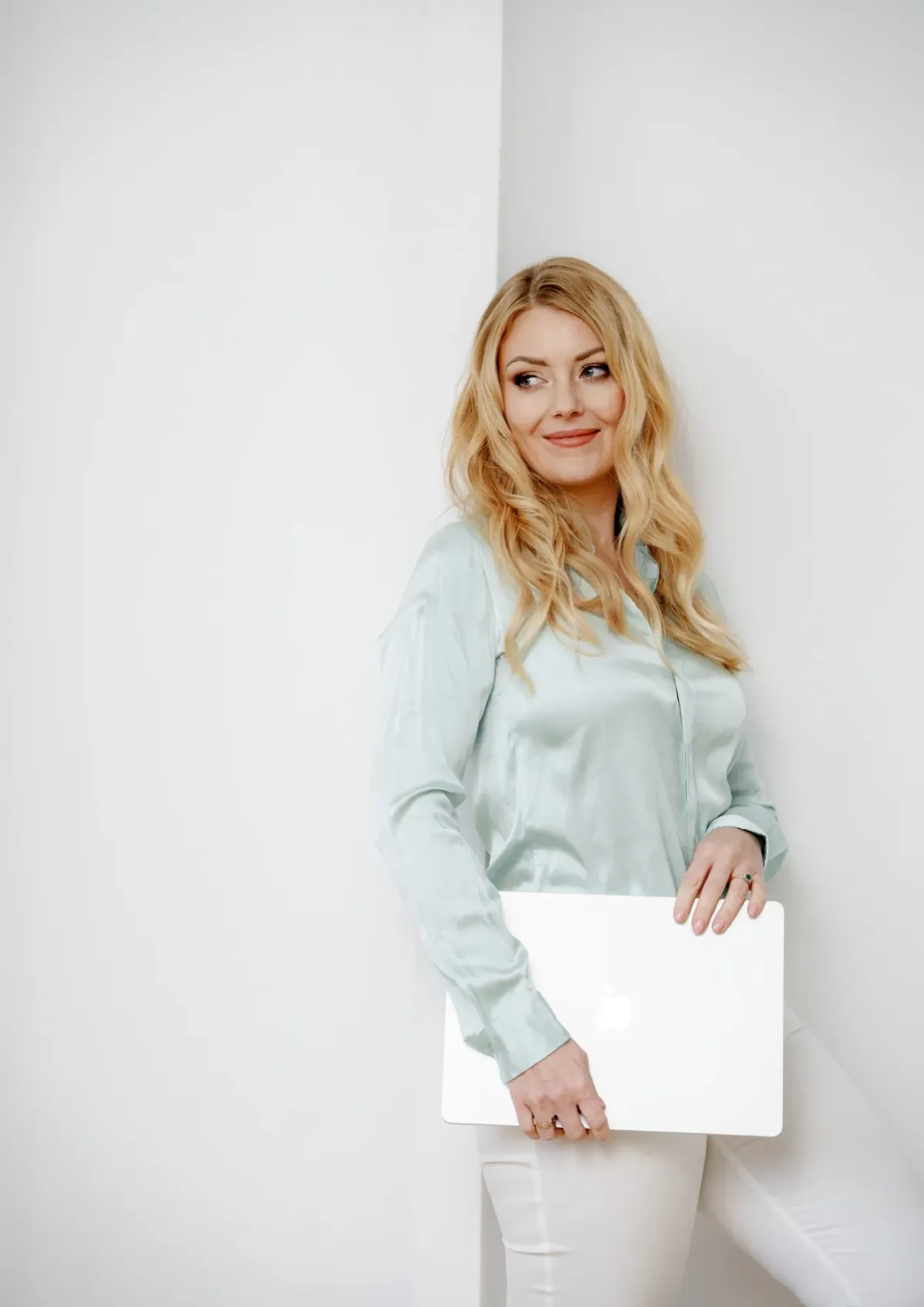 Smiling blonde woman in a light blue satin blouse holding a closed MacBook. She’s leaning casually against a white wall, looking to the side with confidence.