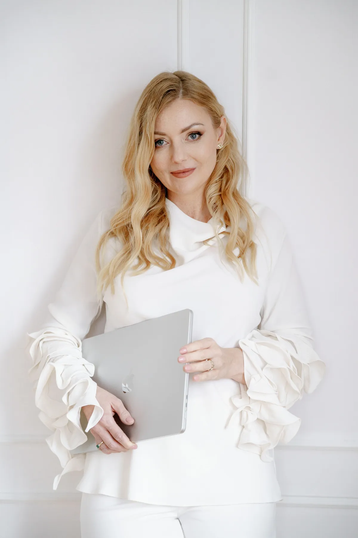 Kobieta w eleganckiej białej bluzce z falbankami, trzymająca srebrnego laptopa, stojąca przy jasnej ścianie. Profesjonalny portret promujący warsztaty i markę Marketing Nattie oraz program The Rising Star.