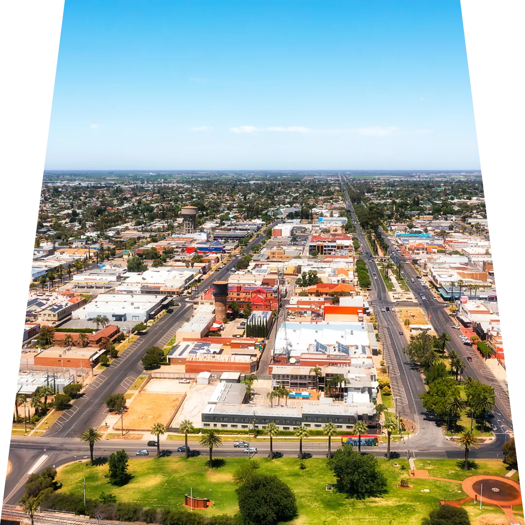  Aerial view of Mildura's central business district and surrounding residential areas, showing the town's grid street layout, mix of commercial and residential buildings, palm tree-lined streets, and expansive rural landscape stretching to the horizon under clear blue skies.