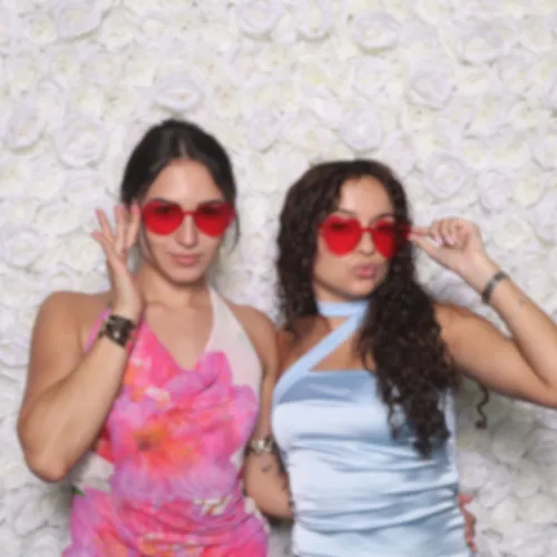 A joyful wedding couple and friends laughing together in front of a MiamiFlicks photo booth, surrounded by lush pastel florals and soft lighting, with elegant wedding attire and a romantic, airy atmosphere.
