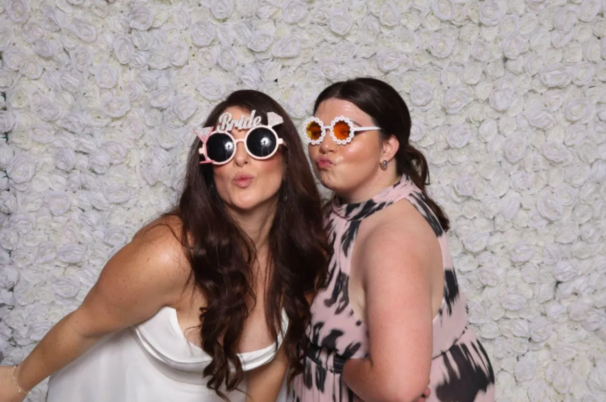 Bride and groom sharing a candid laugh in front of a MiamiFlicks booth, with a lush green wall and gold accents, surrounded by friends in pastel dresses and suits.