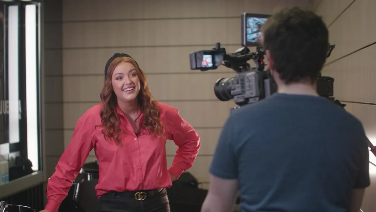 Camera operator filming a presenter in a pink shirt indoors