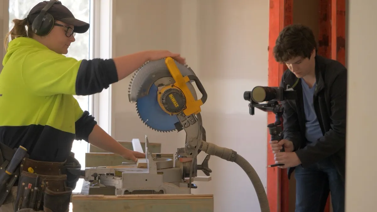 Camera operator filming a worker using a mitre saw inside a building