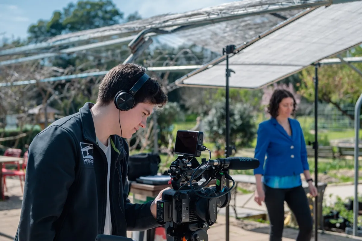 Camera operator checking the rig while the interview participant waits among plants