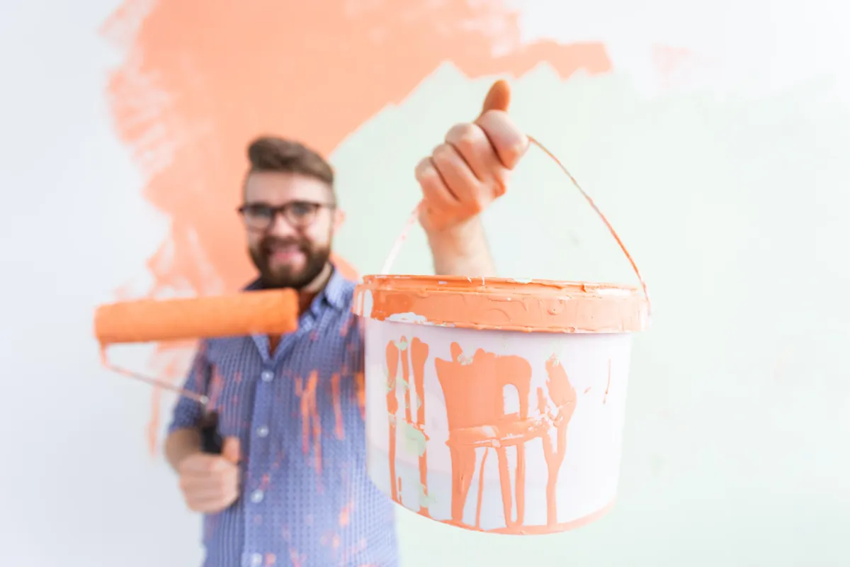 a person holding a paint roller and a bucket of orange paint