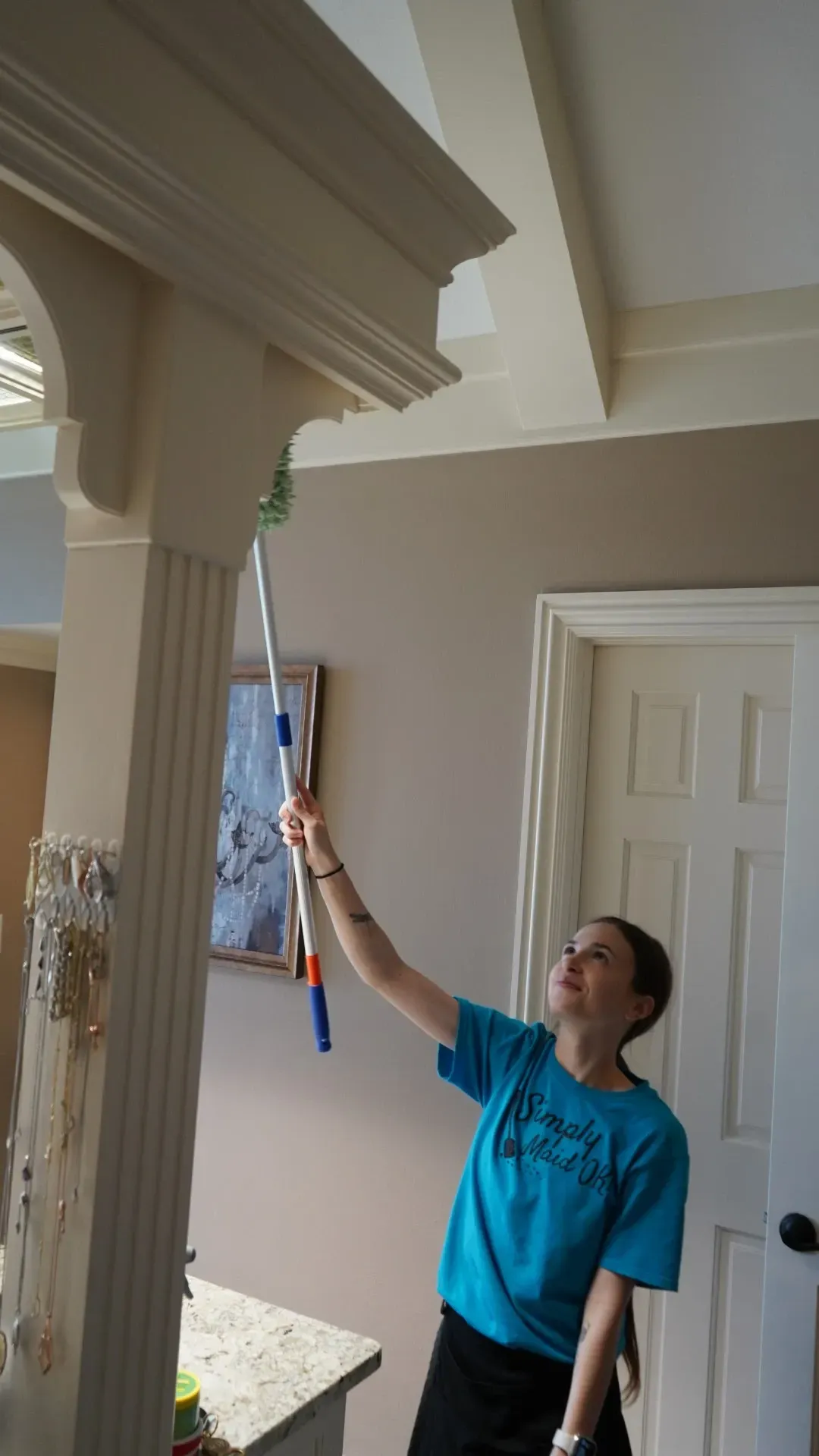Woman in a blue Simply Maid t-shirt using a long duster to clean high corners in a home, showcasing personalized cleaning services in Skiatook, Oklahoma.