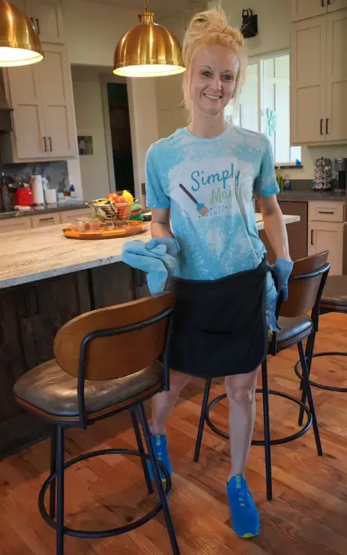 Woman in a Simply Maid t-shirt and apron, holding cleaning cloths, standing in a modern kitchen with bar stools and a wooden floor, representing the professional cleaning services offered by Simply Maid.