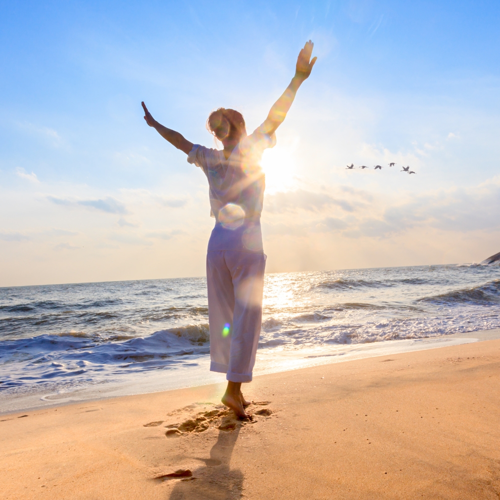 A woman spreading her arms on the beach with a flock of birds flying in the distance
