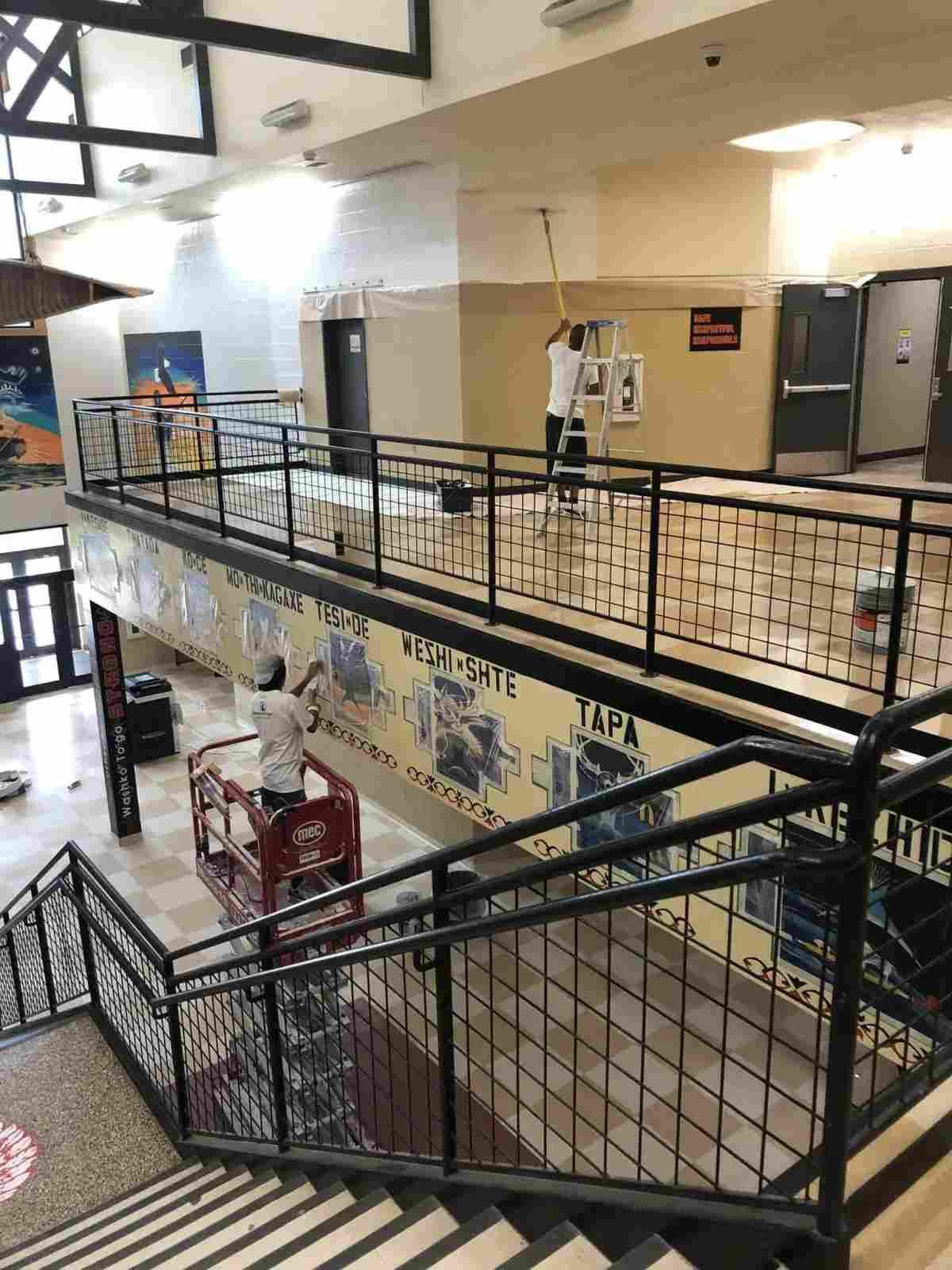 Two workers painting the walls of a school lobby; one on a ladder and one on a lift, surrounded by murals and a staircase.