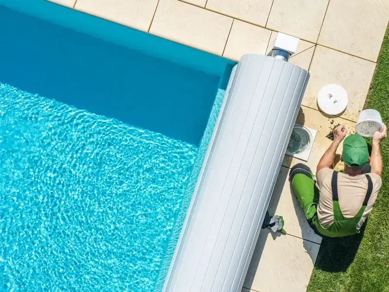 Overhead view of a pool inspector checking the filter basket beside a covered pool.