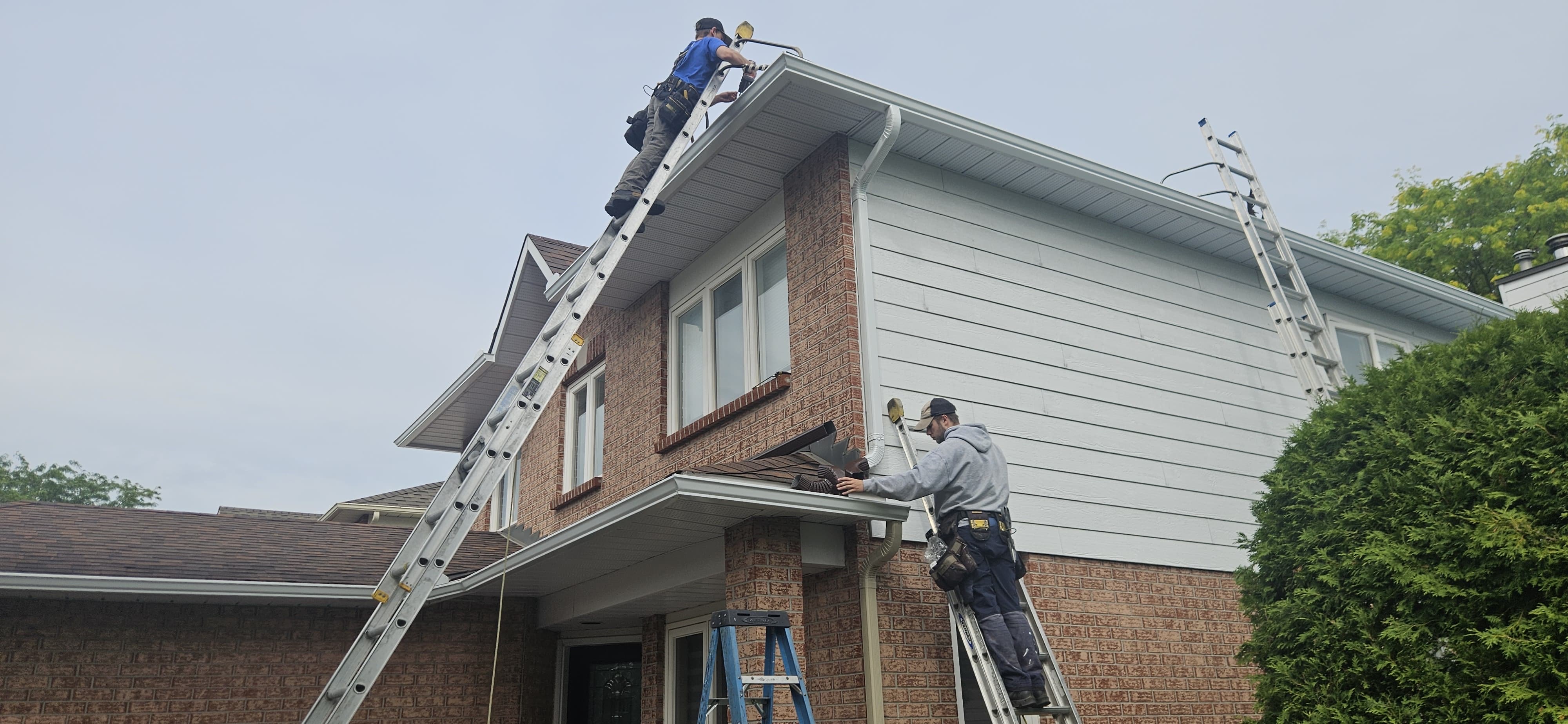 Professional roofing crew using extension ladders to install white gutters and downspouts on a two-story brick home.