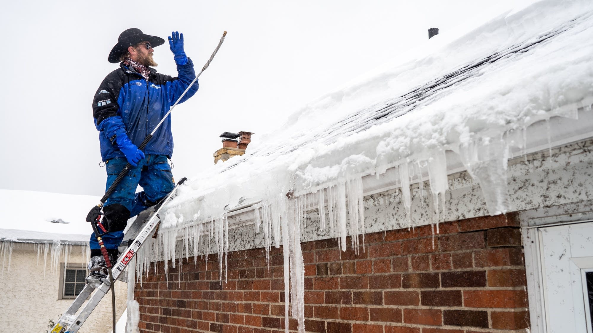 Professional contractor on a ladder preparing to remove heavy ice dams and icicles from a brick house roof.
