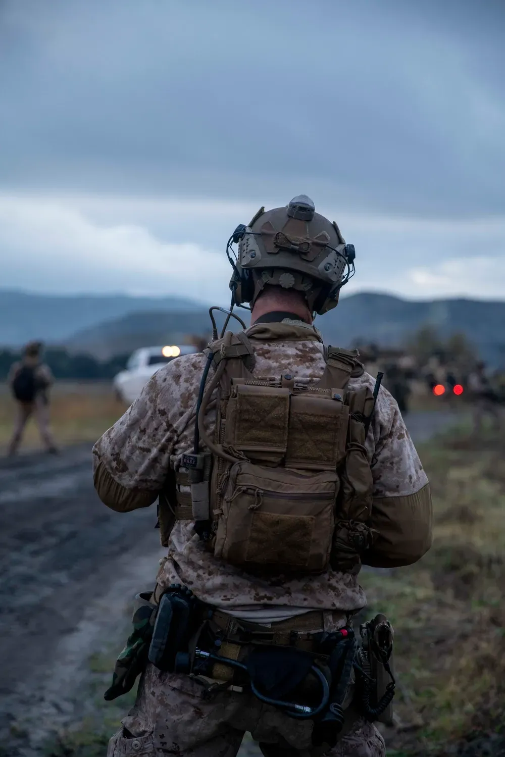 A soldier in full gear walks along a rural road at dusk, mountains visible in the distance. The mood is tense and atmospheric.