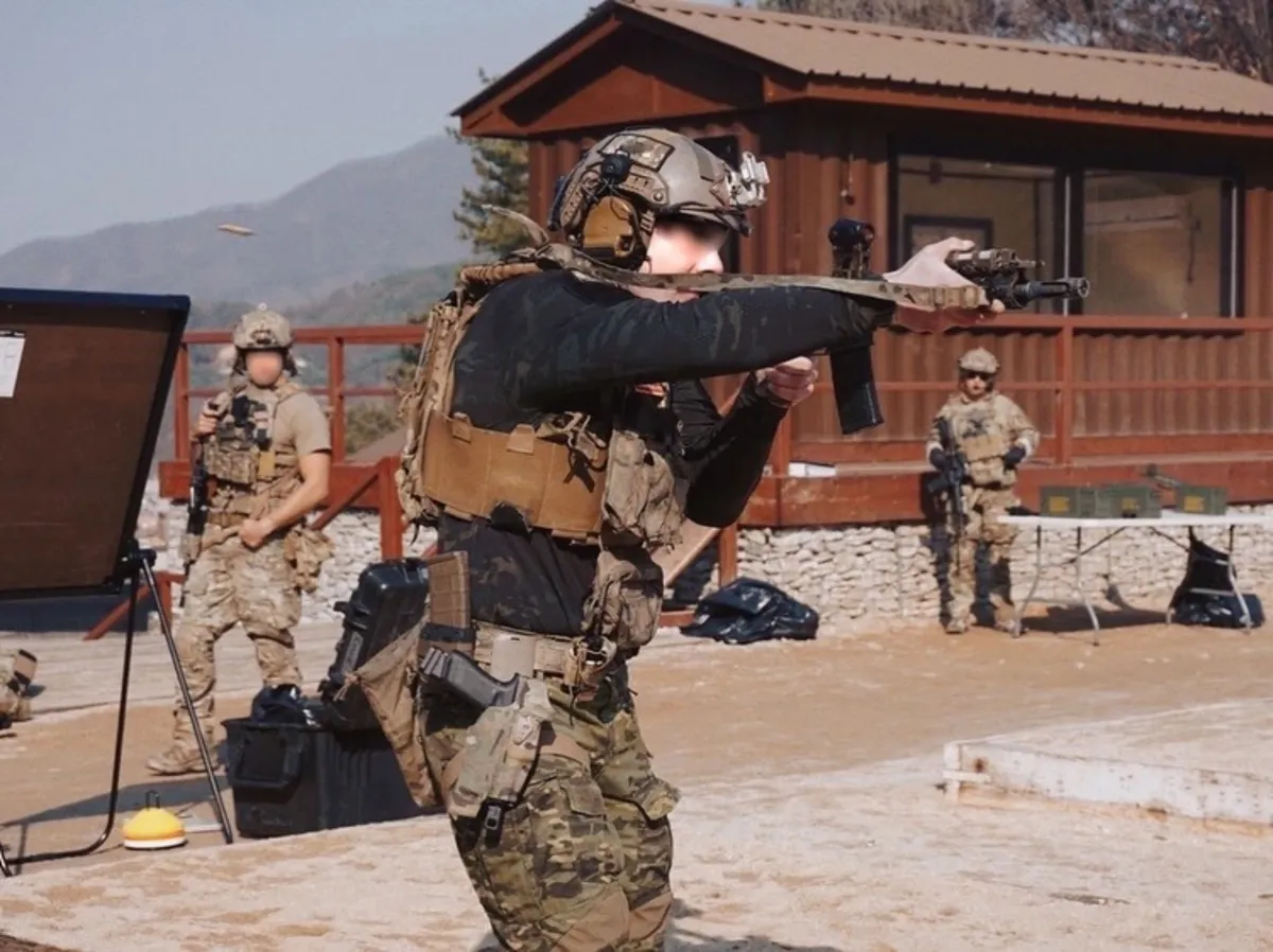 A soldier in full tactical gear trains outside, aiming a handgun. Two other soldiers observe, with a wooden building and mountains in the background.