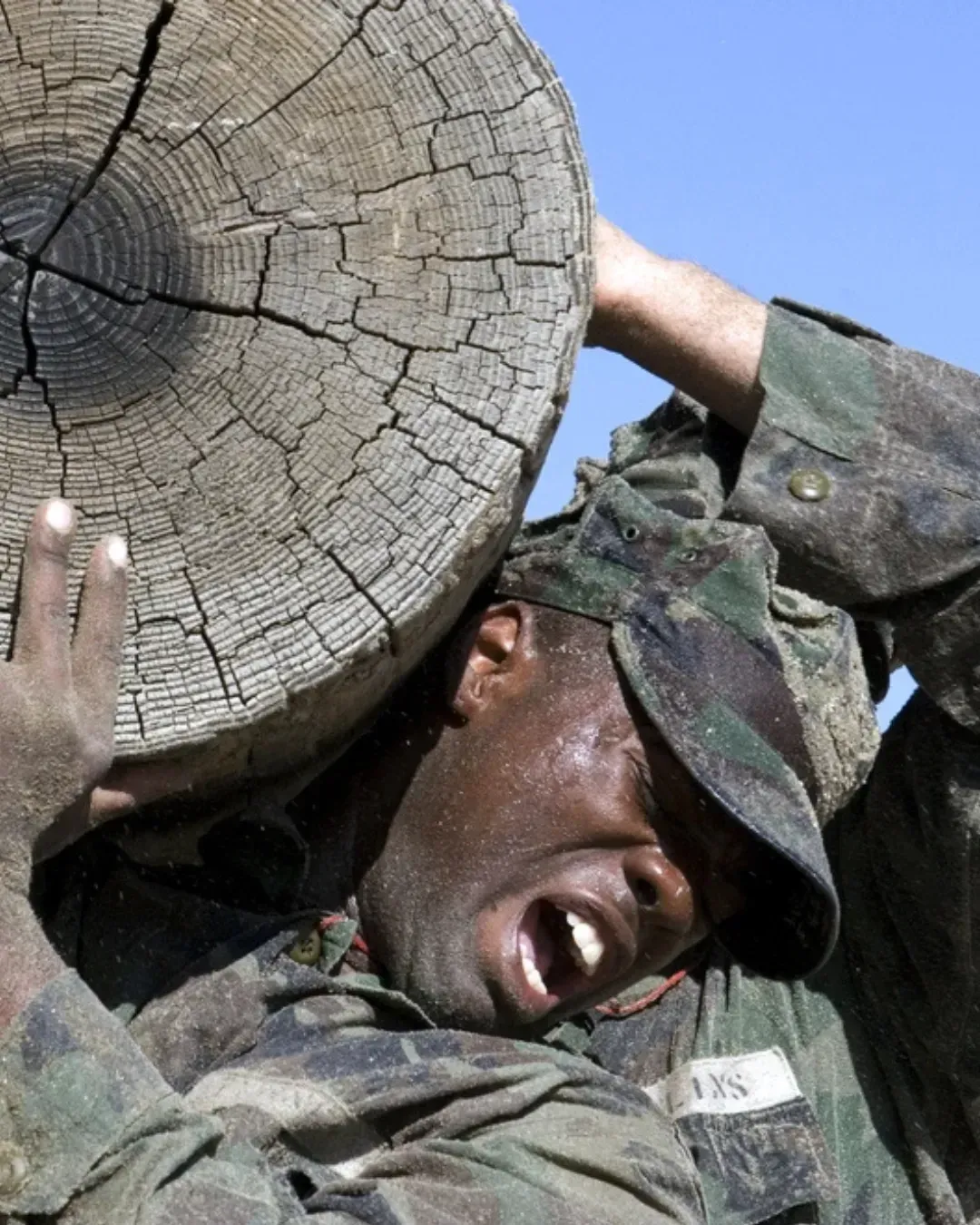 A soldier in camouflage struggles to carry a heavy log during training. His expression shows exertion under a clear blue sky, conveying determination and endurance.