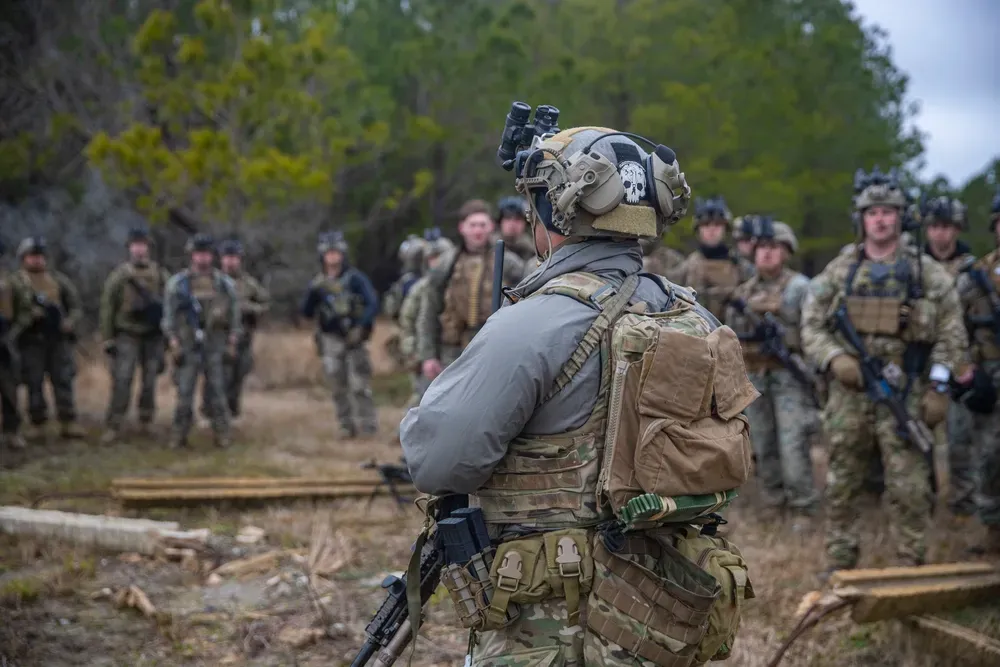 A group of soldiers in camouflage gear stand in a forest clearing. They appear attentive and focused, suggesting a training exercise or briefing.