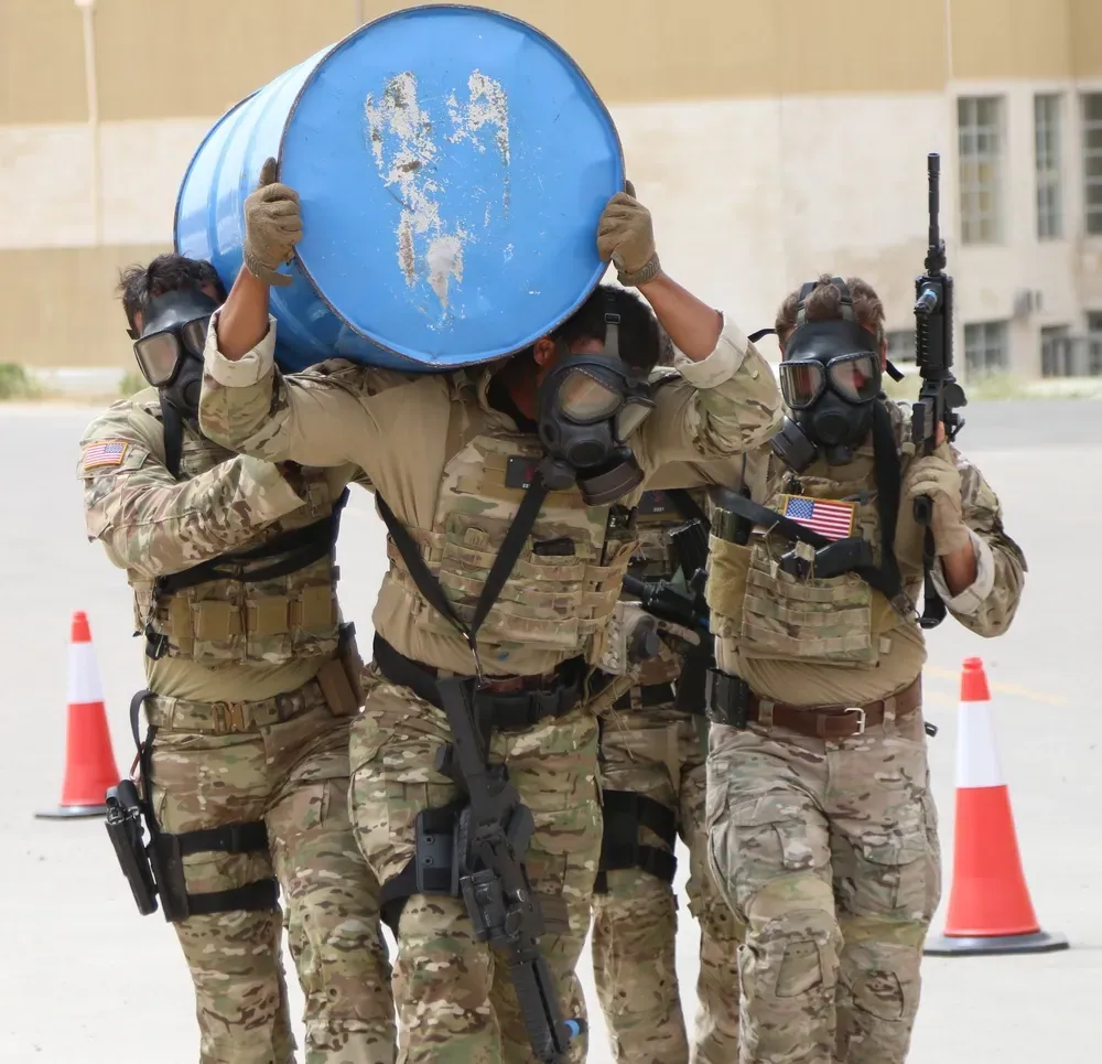 Four soldiers in camouflage and helmets, wearing gas masks, carry a blue barrel in an urban training setting. Orange cones are visible in the background.
