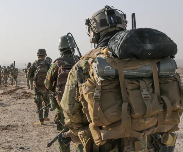 Soldiers in military gear march in a desert landscape, carrying backpacks. The scene conveys discipline and focus under a clear, bright sky.