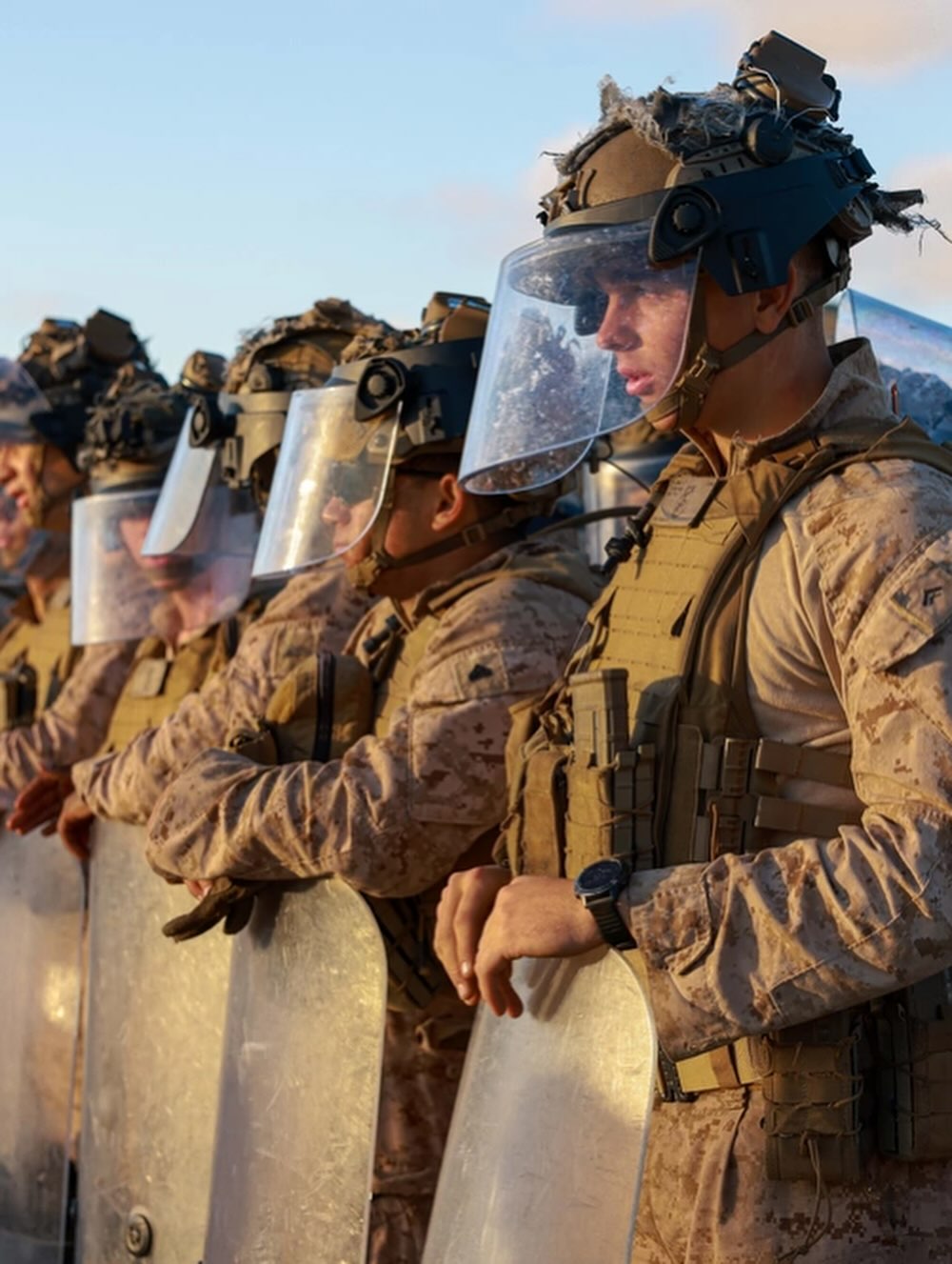 Soldiers in tactical gear and helmets stand in line, holding transparent shields. The scene conveys readiness and discipline under a clear sky.