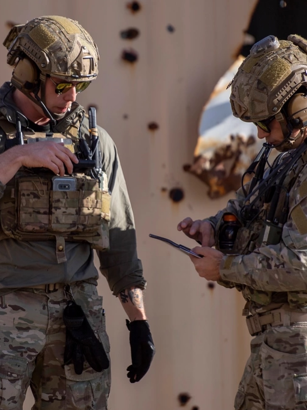 Two soldiers in tactical gear stand beside each other, focusing on a device one is holding. Their expressions convey concentration and seriousness.