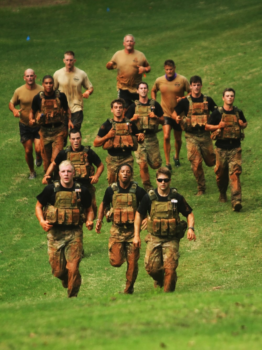A group of eleven people runs uphill on a grassy field, all wearing tactical gear and camouflage uniforms, conveying teamwork and determination.