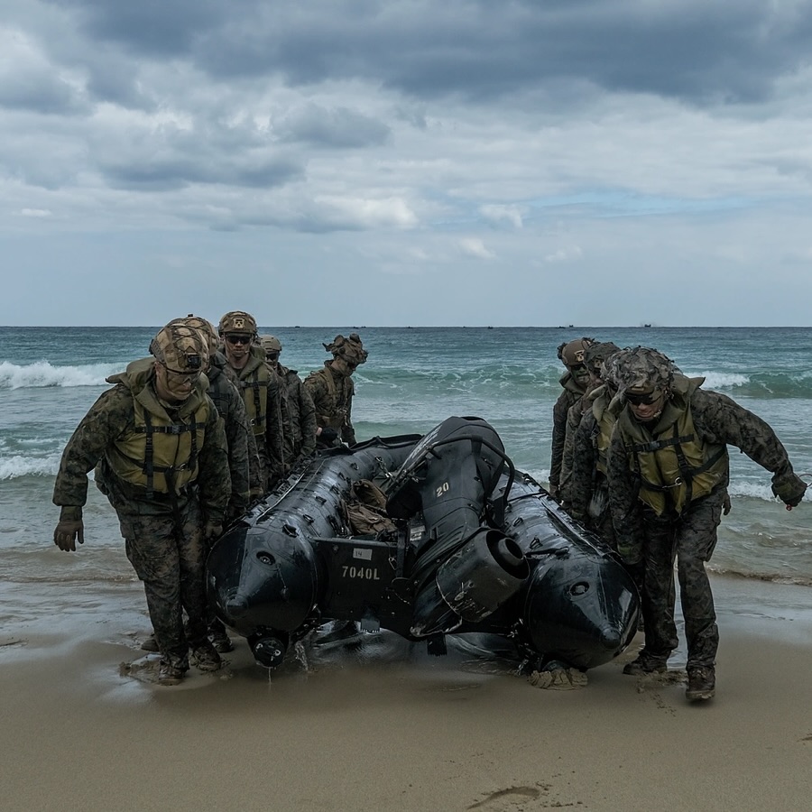 Soldiers in camouflage carry an inflatable boat ashore under an overcast sky. The ocean waves and cloudy weather set a tense, determined mood.