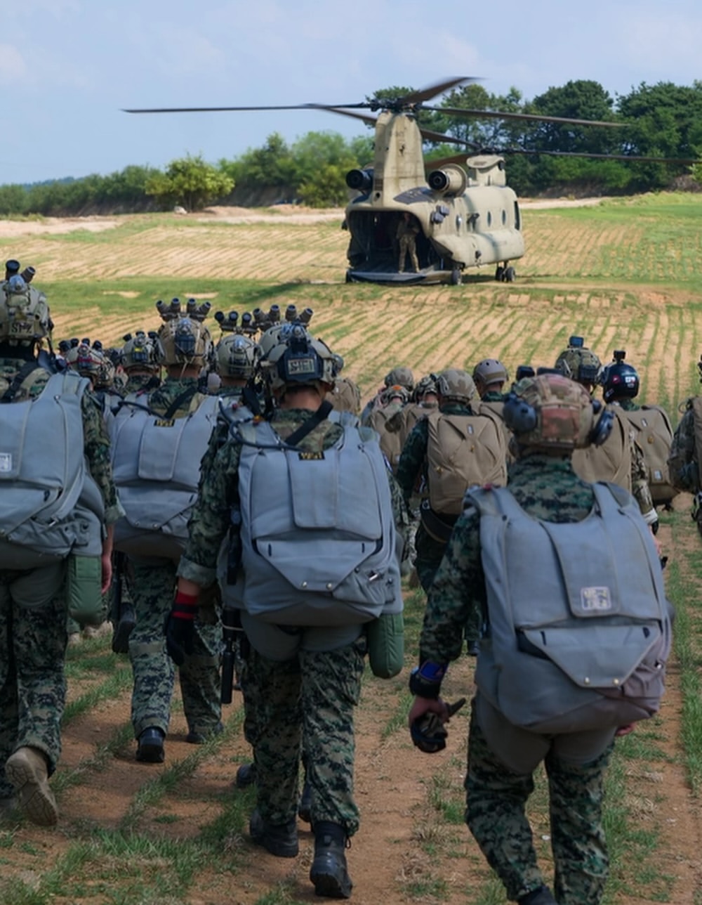 Soldiers in uniform with backpacks approach a large helicopter on a grassy field. The scene conveys a sense of urgency and coordination.