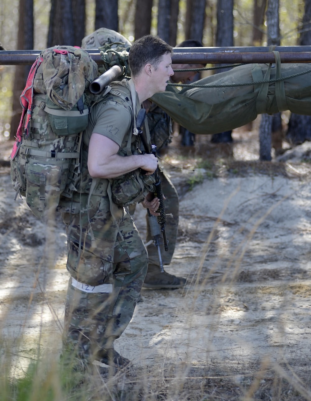 A soldier in camouflage carries a heavy backpack, pulling a stretcher in a forest setting. The image conveys determination and teamwork.