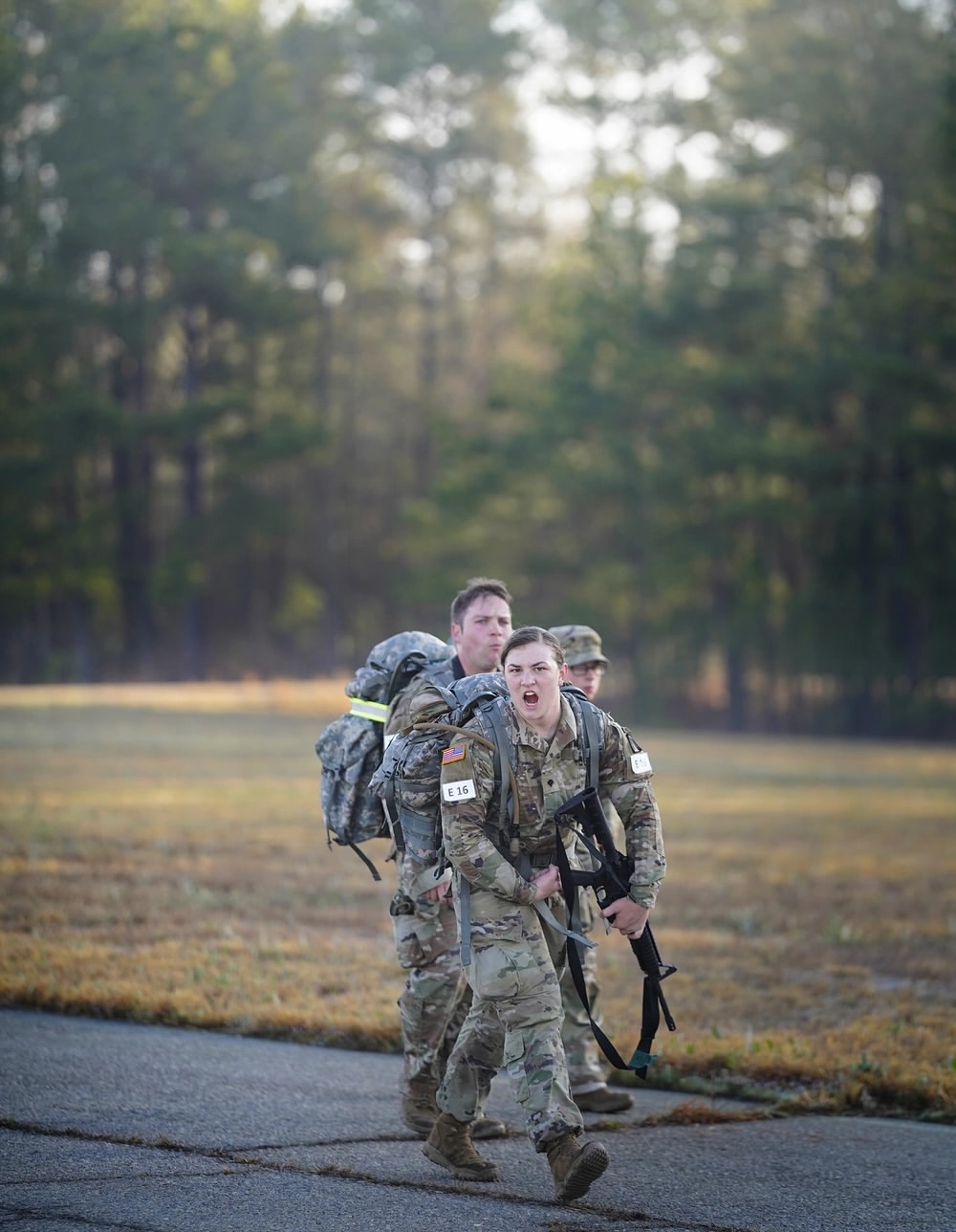 Three soldiers in camouflage uniforms carry heavy backpacks while running on a path through a forested area, conveying determination and teamwork.
