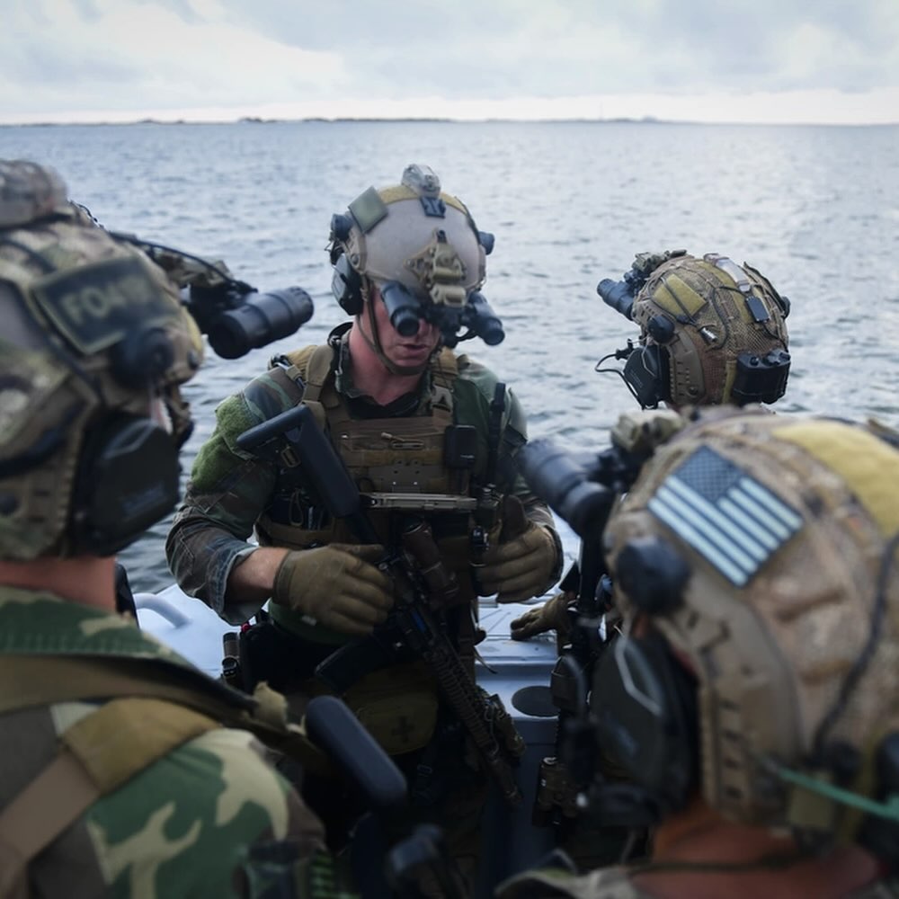 Four soldiers in camouflage gear and helmets with night vision goggles preparing for a mission on a boat, set against a calm sea background.