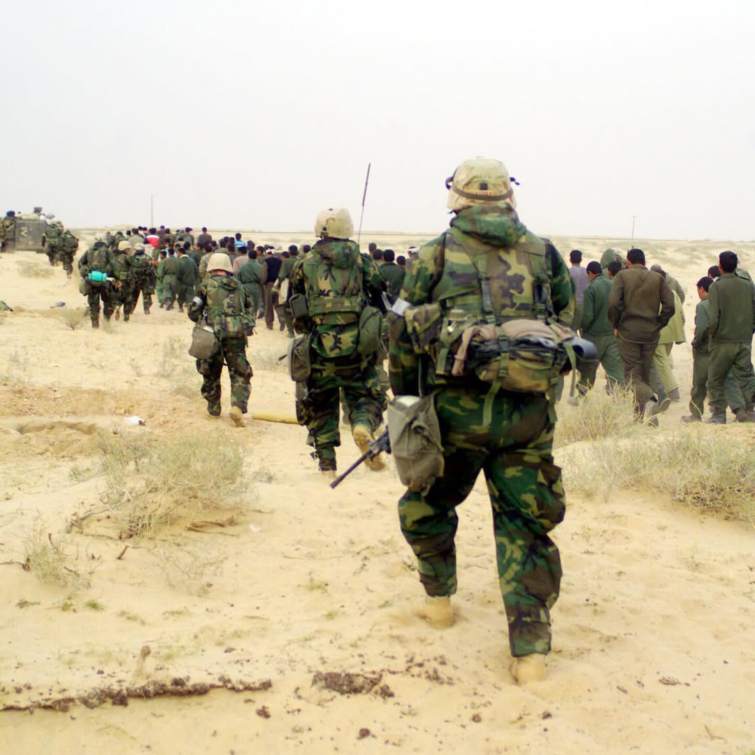 Soldiers in camouflage walk through a desert landscape, leading a group of civilians. The atmosphere is tense and orderly, with a cloudy sky overhead.