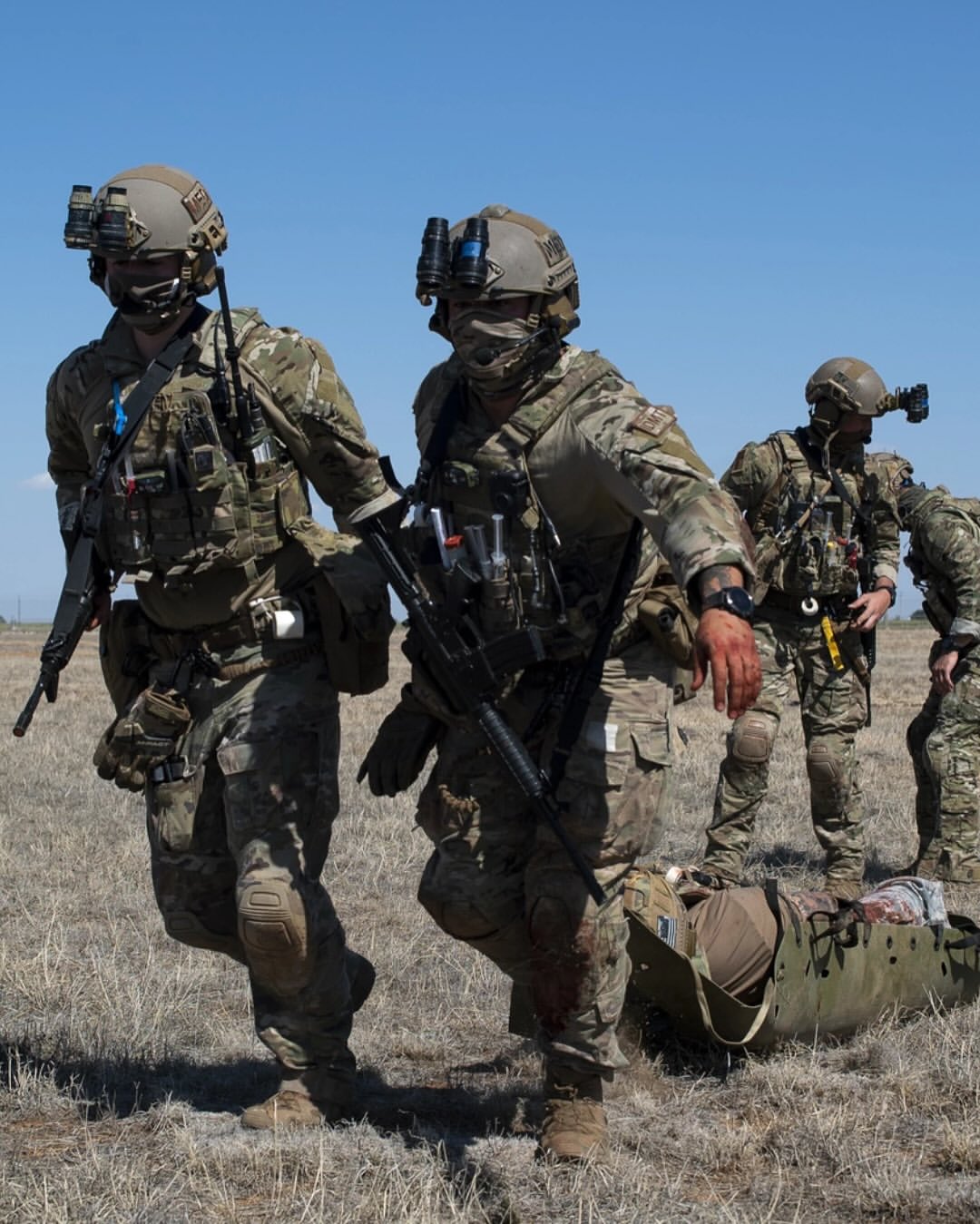 Soldiers in camouflage and helmets run across a dry field with military gear, conveying urgency and teamwork under a clear blue sky.