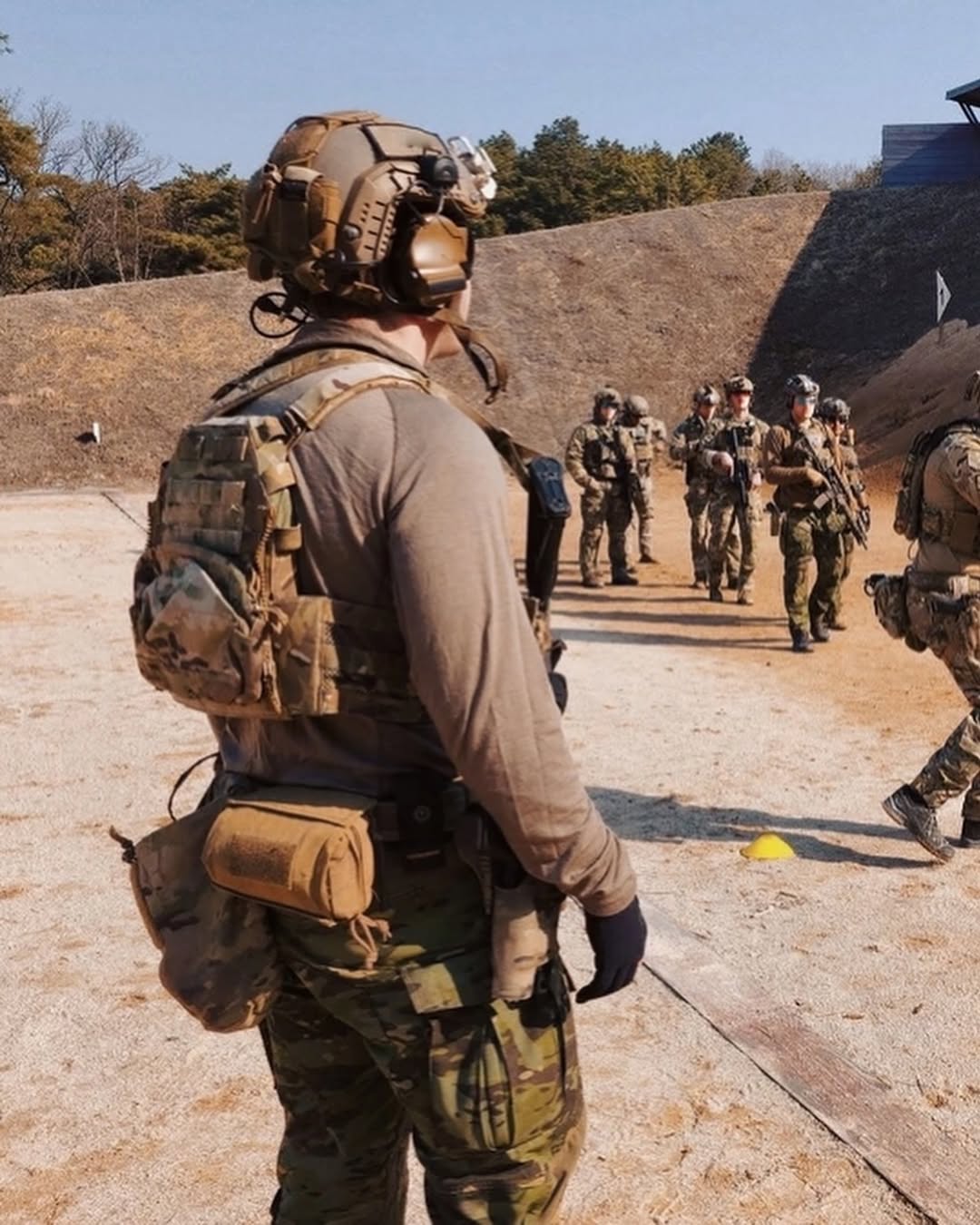 A group of soldiers in tactical gear stand on a sandy training ground. One in the foreground carries a backpack, facing others who are gathered in the background.